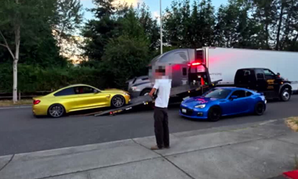 Yellow sports car and blue sports car are being loaded onto a tow trailer on a residential street, with a man standing nearby and a large white trailer truck behind them.