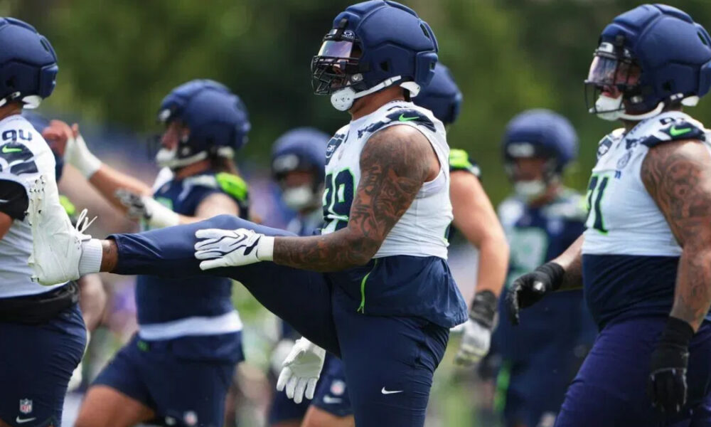 American football players in navy and white practice uniforms stretch on a grassy field, wearing helmets and gloves.