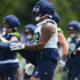 American football players in navy and white practice uniforms stretch on a grassy field, wearing helmets and gloves.