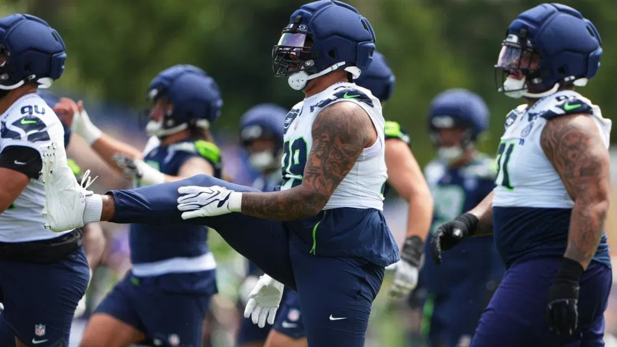 American football players in navy and white practice uniforms stretch on a grassy field, wearing helmets and gloves.