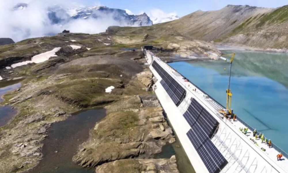 Dam under construction with solar panels along its side crossing a turquoise alpine lake in a rocky mountain landscape with a crane and workers on site.