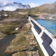 Dam under construction with solar panels along its side crossing a turquoise alpine lake in a rocky mountain landscape with a crane and workers on site.