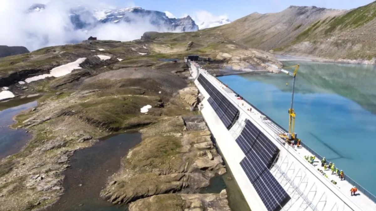 Dam under construction with solar panels along its side crossing a turquoise alpine lake in a rocky mountain landscape with a crane and workers on site.