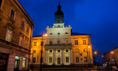 Night view of a neoclassical town hall glowing yellow, with a blue evening sky and street lamps.