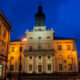 Night view of a neoclassical town hall glowing yellow, with a blue evening sky and street lamps.