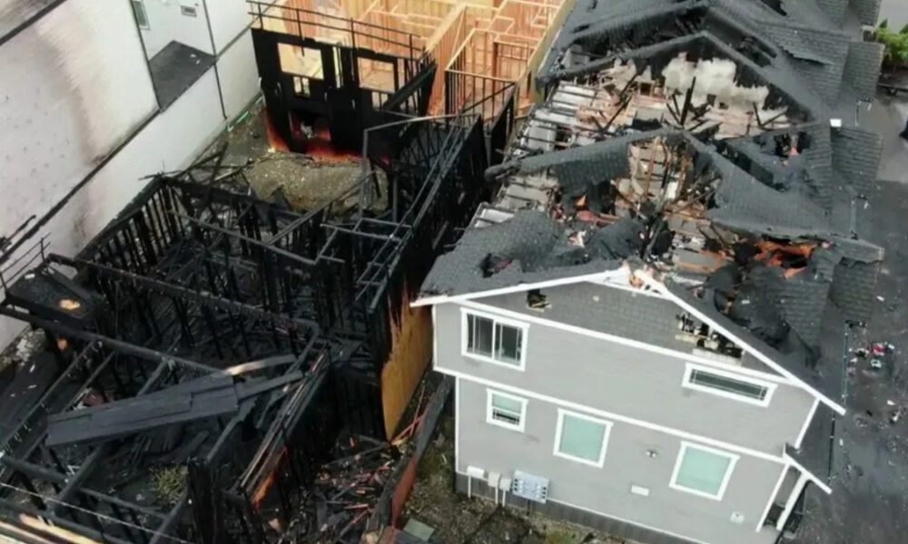 Gray two-story house in foreground, with a heavily damaged, fire-devastated structure and charred framing behind it. The roof is collapsed and debris is scattered around the site.
