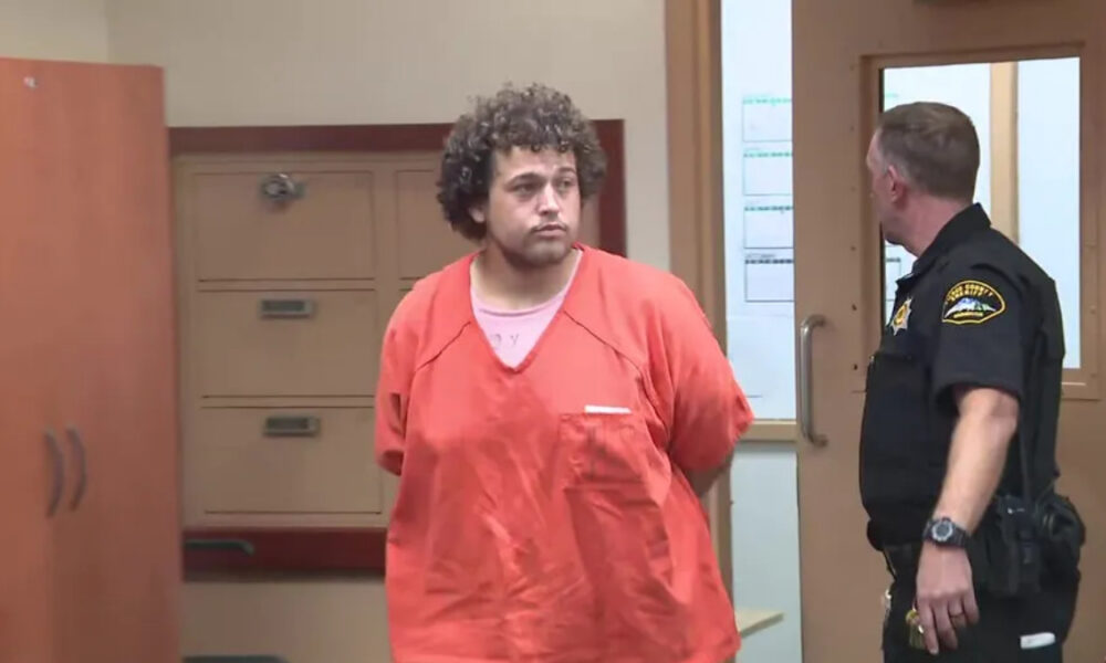 Man in an orange jail jumpsuit stands with hands behind back as a police officer in a dark uniform speaks to him in a hallway.
