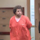 Man in an orange jail jumpsuit stands with hands behind back as a police officer in a dark uniform speaks to him in a hallway.