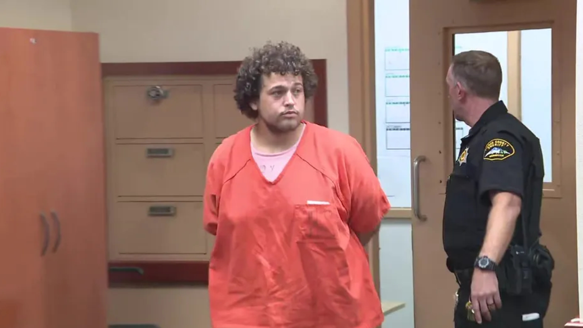 Man in an orange jail jumpsuit stands with hands behind back as a police officer in a dark uniform speaks to him in a hallway.