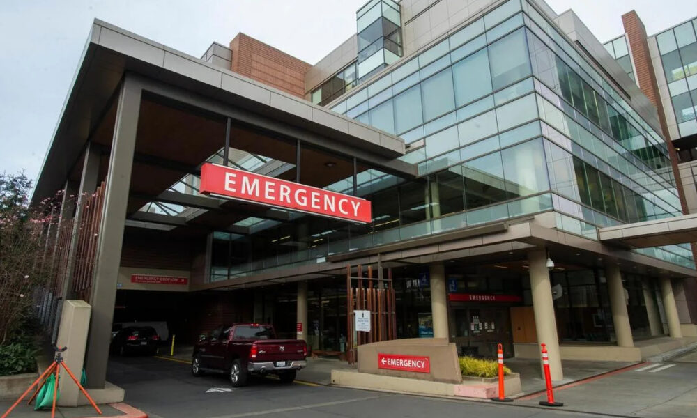 Exterior view of a hospital emergency department entrance with a prominent red EMERGENCY sign and vehicles in the driveway