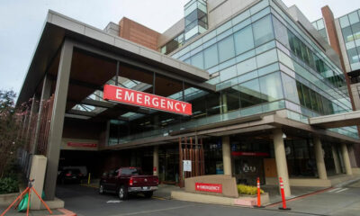 Exterior view of a hospital emergency department entrance with a prominent red EMERGENCY sign and vehicles in the driveway