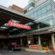 Exterior view of a hospital emergency department entrance with a prominent red EMERGENCY sign and vehicles in the driveway