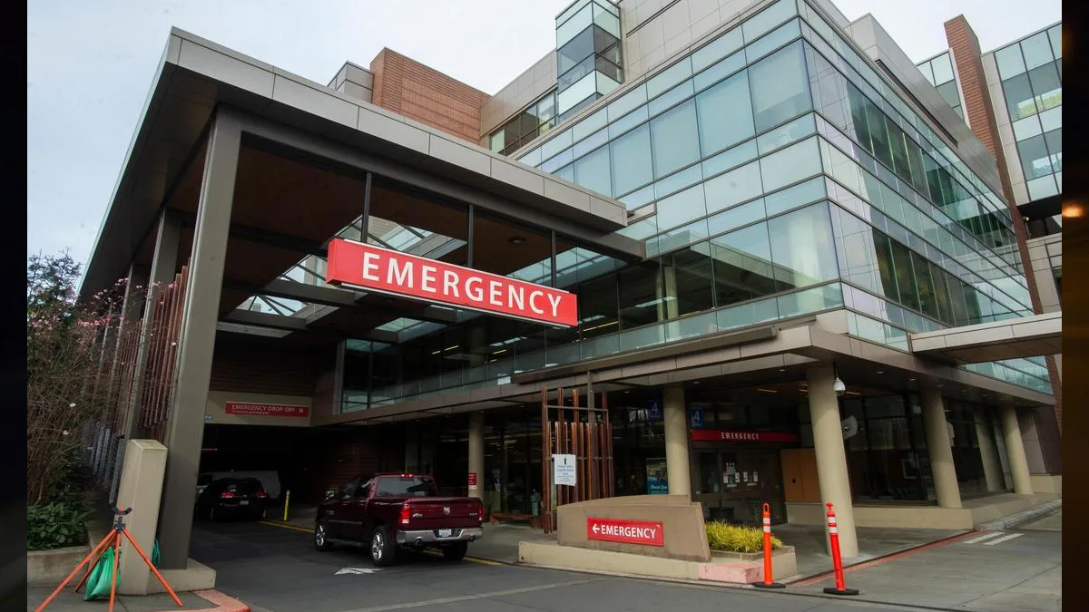 Exterior view of a hospital emergency department entrance with a prominent red EMERGENCY sign and vehicles in the driveway
