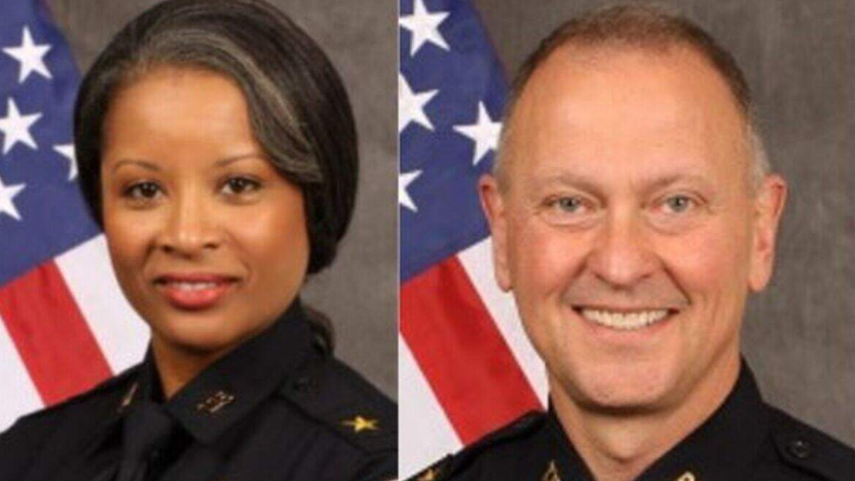 Two police officers, a woman (left) and a man (right), in formal uniform with an American flag backdrop.