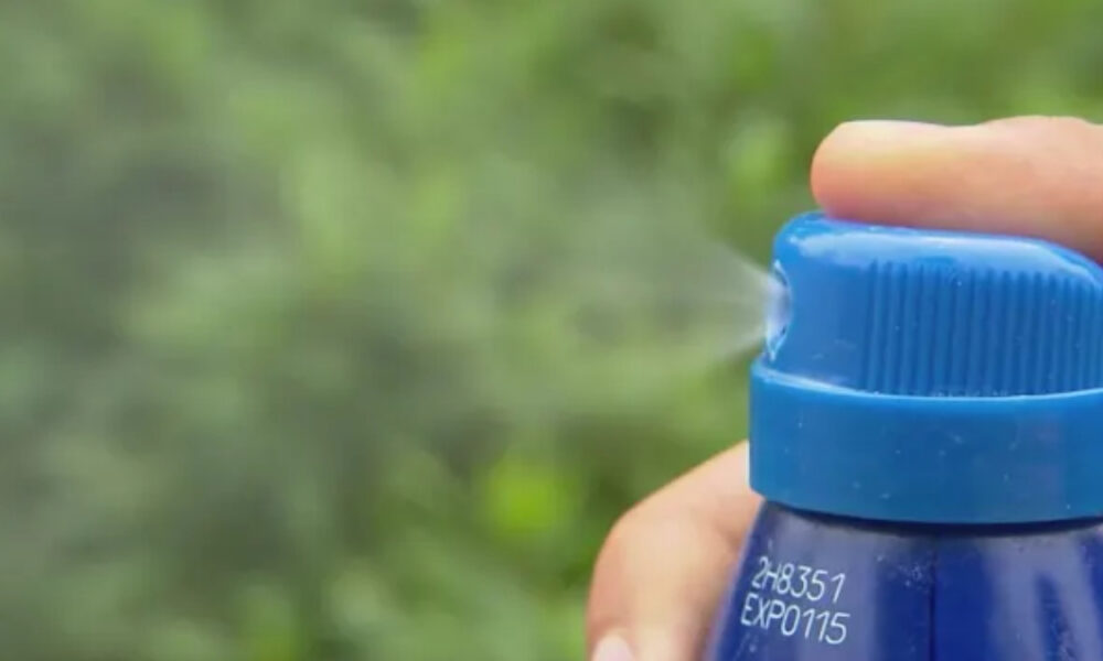 Close-up of a blue spray bottle releasing a fine mist outdoors with blurred green foliage in the background, held by a hand.