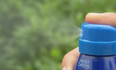 Close-up of a blue spray bottle releasing a fine mist outdoors with blurred green foliage in the background, held by a hand.