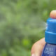 Close-up of a blue spray bottle releasing a fine mist outdoors with blurred green foliage in the background, held by a hand.
