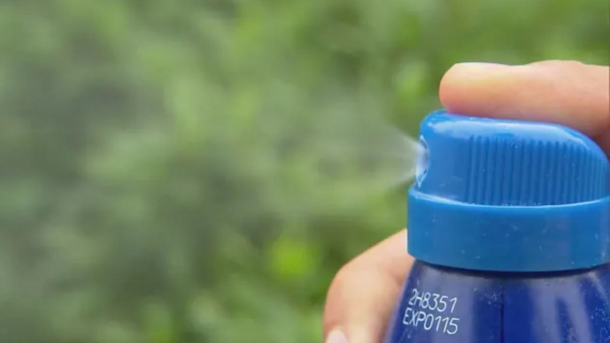 Close-up of a blue spray bottle releasing a fine mist outdoors with blurred green foliage in the background, held by a hand.