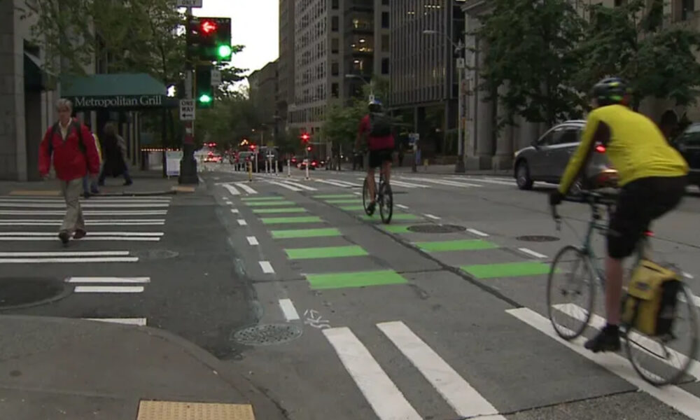Urban street scene with cyclists riding in a bike lane and pedestrians crossing at a crosswalk near a metropolitan building and cafe sign.
