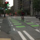 Urban street scene with cyclists riding in a bike lane and pedestrians crossing at a crosswalk near a metropolitan building and cafe sign.