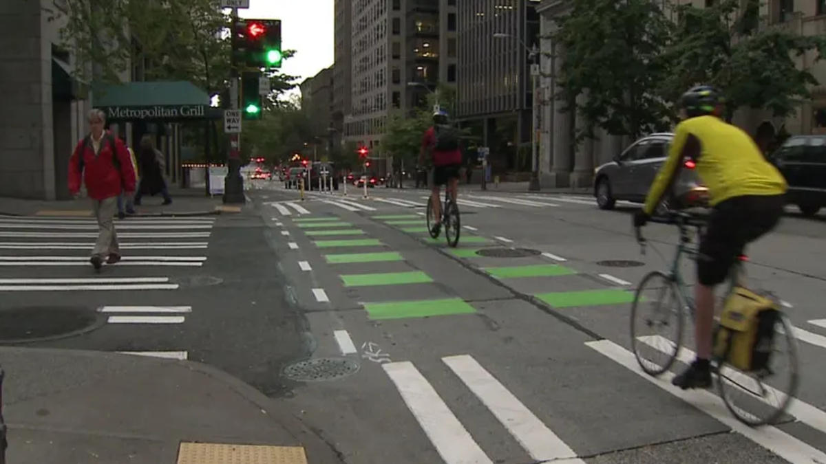 Urban street scene with cyclists riding in a bike lane and pedestrians crossing at a crosswalk near a metropolitan building and cafe sign.