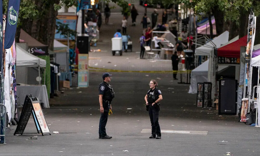 Two police officers stand facing each other on a closed street at a festival, with vendor tents and yellow tape in the background.