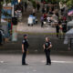 Two police officers stand facing each other on a closed street at a festival, with vendor tents and yellow tape in the background.