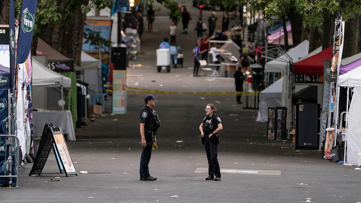 Two police officers stand facing each other on a closed street at a festival, with vendor tents and yellow tape in the background.