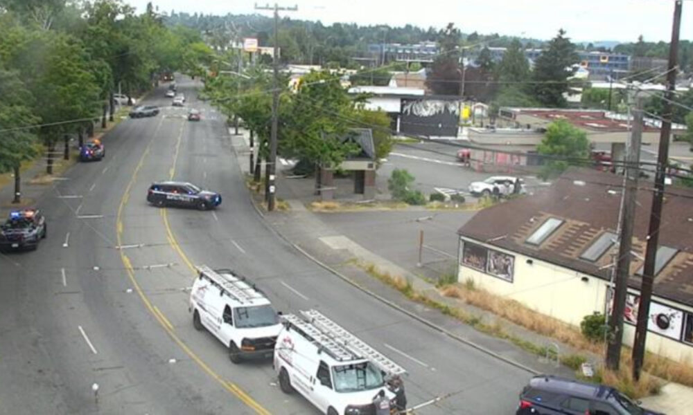 Police cars block a curved city street while white service vans with ladders park nearby; few cars navigate around the scene with buildings and trees in the background.