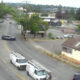 Police cars block a curved city street while white service vans with ladders park nearby; few cars navigate around the scene with buildings and trees in the background.
