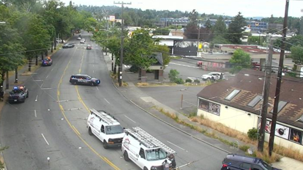 Police cars block a curved city street while white service vans with ladders park nearby; few cars navigate around the scene with buildings and trees in the background.