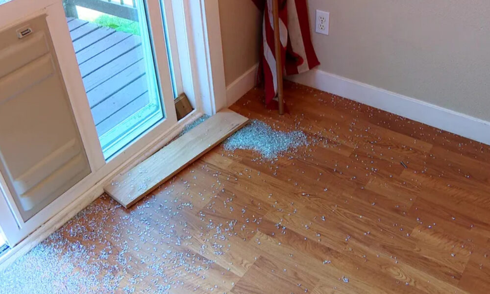 Shattered glass scattered on a hardwood floor by a sliding glass door, with a wooden board resting across the threshold.