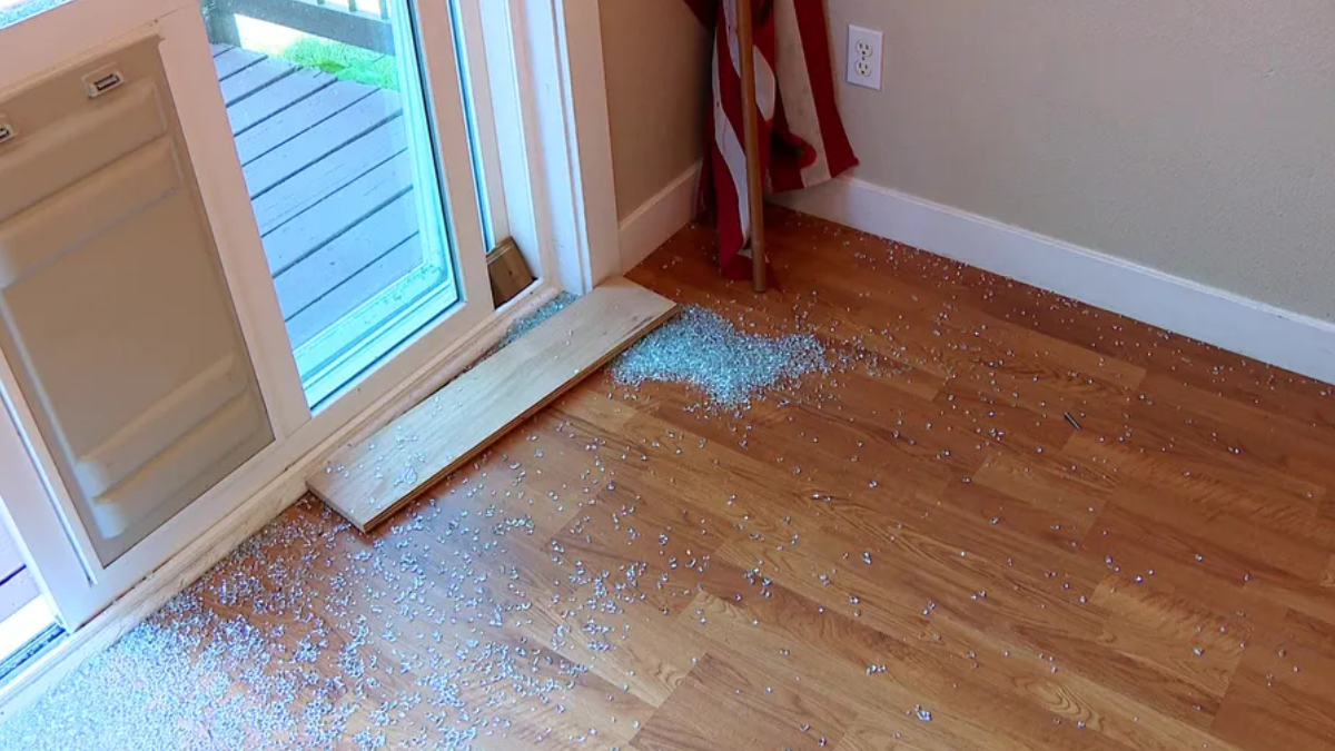 Shattered glass scattered on a hardwood floor by a sliding glass door, with a wooden board resting across the threshold.