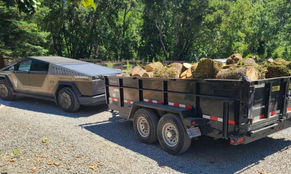 Cybertruck-style pickup towing a black log-filled trailer on a gravel driveway with trees in the background.