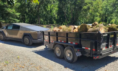Cybertruck-style pickup towing a black log-filled trailer on a gravel driveway with trees in the background.