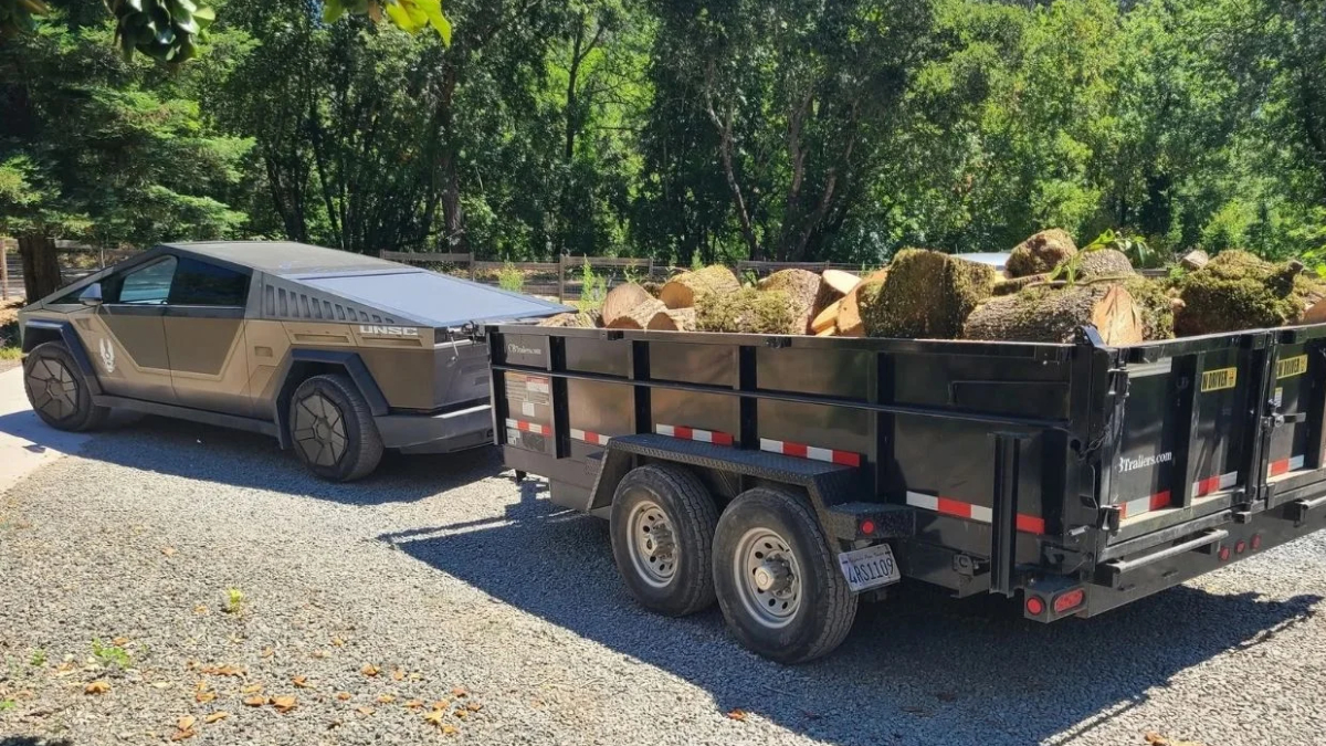 Cybertruck-style pickup towing a black log-filled trailer on a gravel driveway with trees in the background.