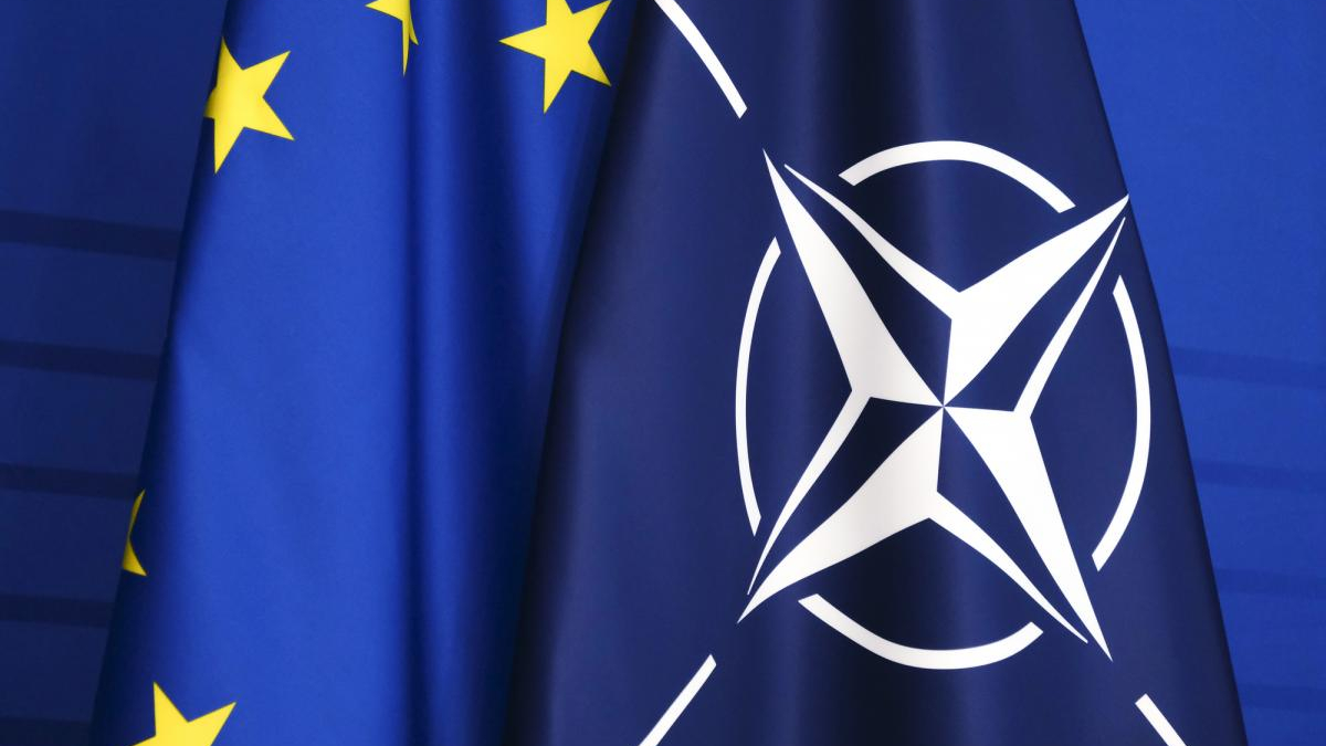 Close-up of blue flags featuring the NATO emblem and partial European Union stars on the left.