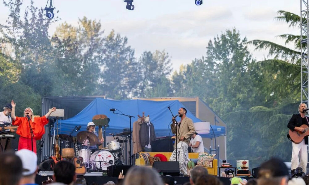 Outdoor concert on a sunny day with a band performing on a stage, singer at mic, guitarist, drummer, and a second vocalist in orange attire.