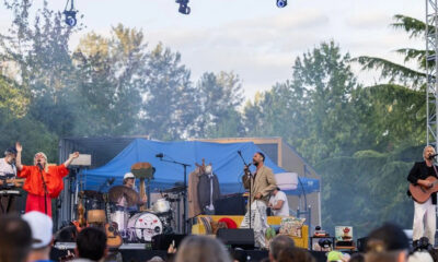 Outdoor concert on a sunny day with a band performing on a stage, singer at mic, guitarist, drummer, and a second vocalist in orange attire.