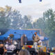Outdoor concert on a sunny day with a band performing on a stage, singer at mic, guitarist, drummer, and a second vocalist in orange attire.