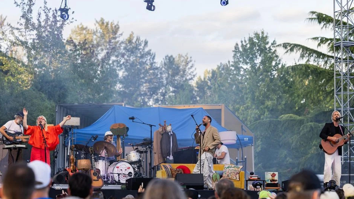 Outdoor concert on a sunny day with a band performing on a stage, singer at mic, guitarist, drummer, and a second vocalist in orange attire.