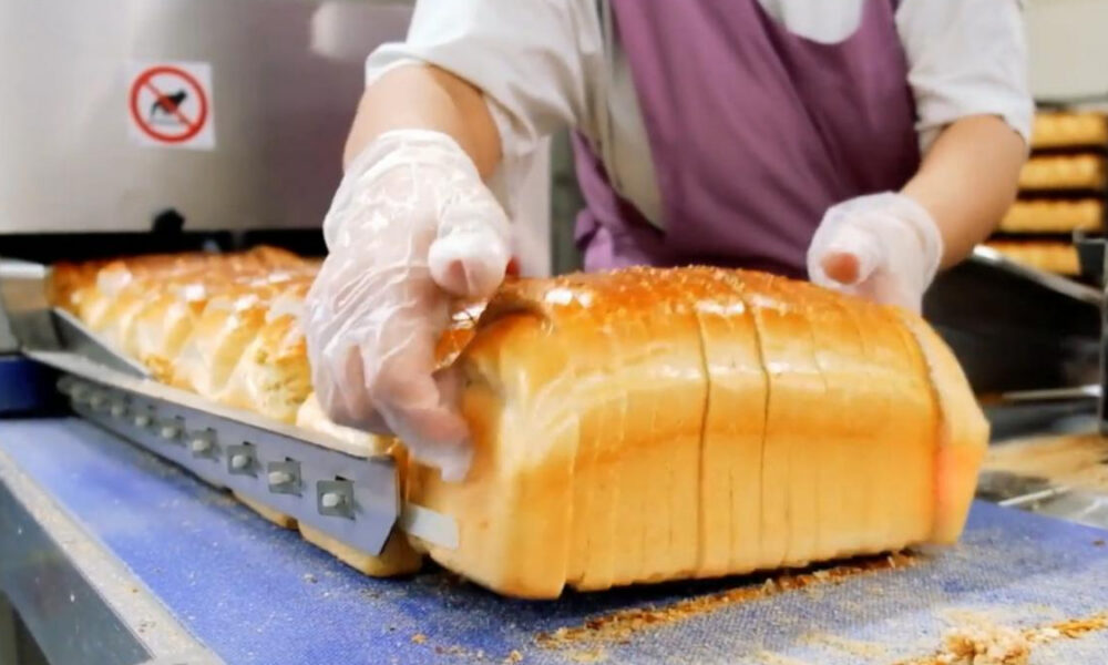A baker wearing gloves slices a large loaf of white bread on a blue conveyor belt in a bakery kitchen.