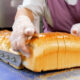 A baker wearing gloves slices a large loaf of white bread on a blue conveyor belt in a bakery kitchen.