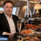 Man in a dark suit sits at a luxurious private-jet dining table with lobster, champagne, and display models of SpaceX and a Tesla car.