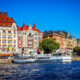 Colorful historic buildings with orange awnings line the harbor as white passenger boats sail by under a blue sky.