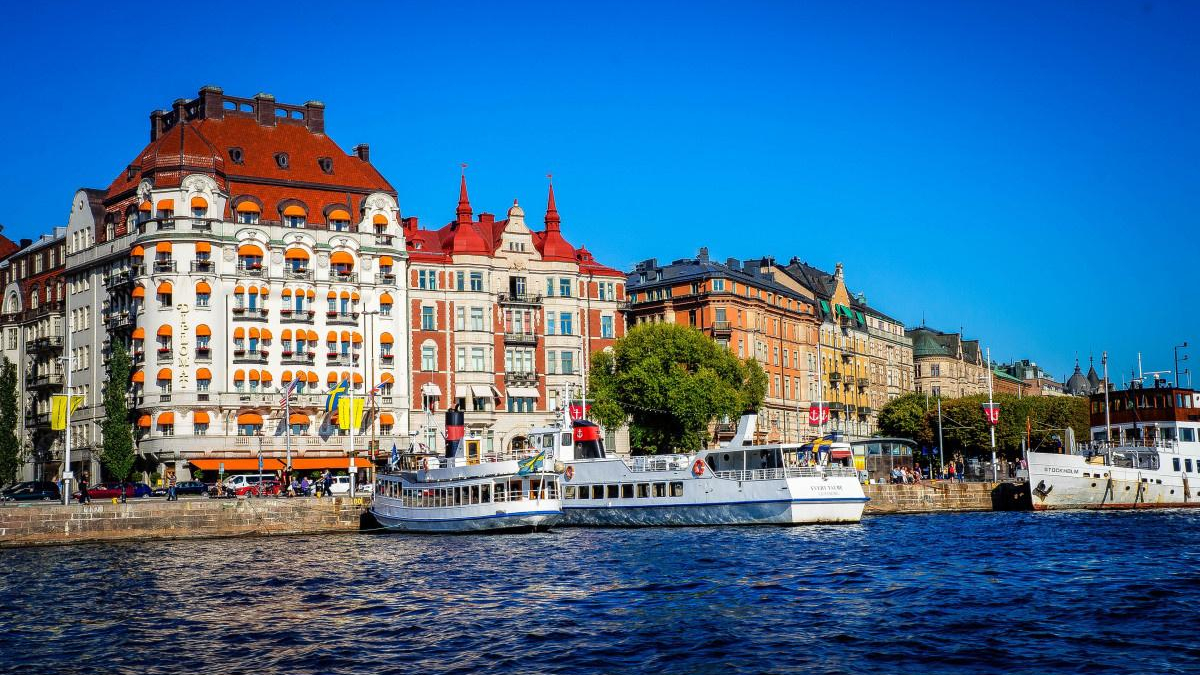 Colorful historic buildings with orange awnings line the harbor as white passenger boats sail by under a blue sky.