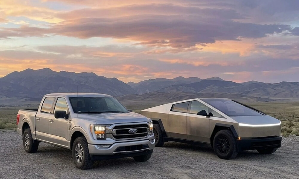 Silver Ford pickup and a metallic Tesla Cybertruck parked on a gravel desert with distant mountains and a colorful sunset behind them.
