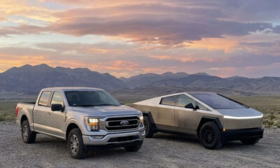 Silver Ford pickup and a metallic Tesla Cybertruck parked on a gravel desert with distant mountains and a colorful sunset behind them.