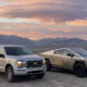 Silver Ford pickup and a metallic Tesla Cybertruck parked on a gravel desert with distant mountains and a colorful sunset behind them.