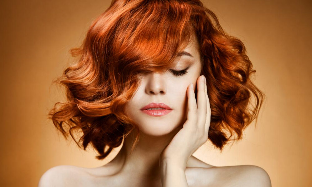 Close-up portrait of a woman with vivid red curly hair, eyes closed, hand resting on her cheek against a warm orange background.
